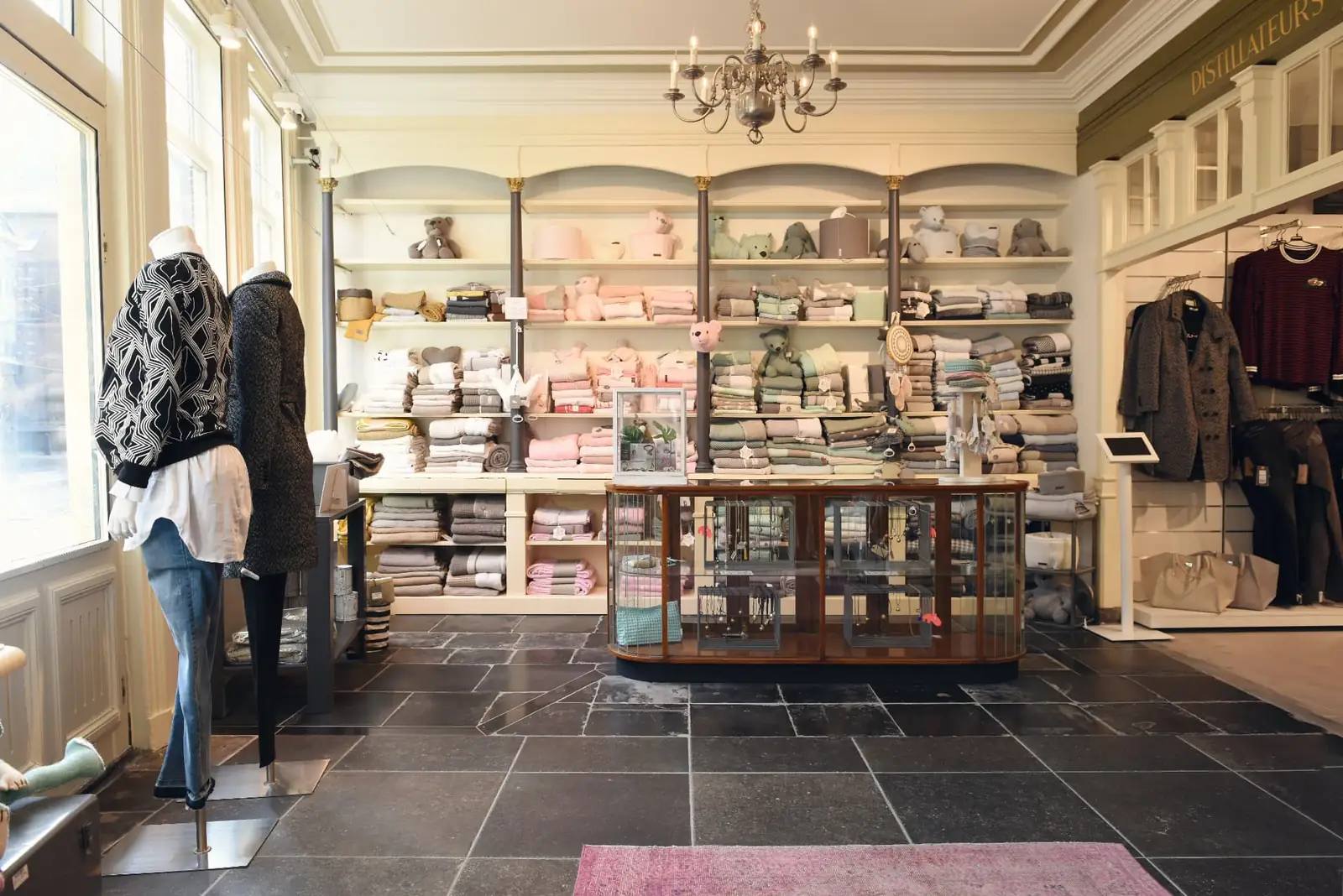 Licht boutique-interieur met een kastenwand vol gevouwen textiel, een toonbank met vitrines en etalagepoppen achter het raam